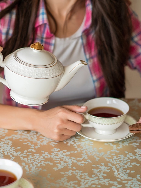 Pouring tea from a porcelain teapot into a cup on a patterned tablecloth.
