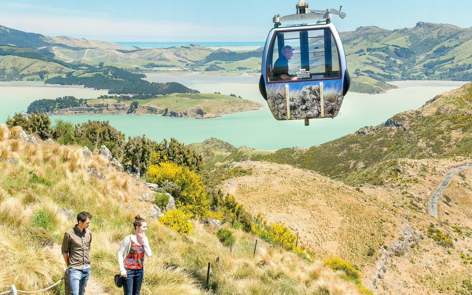 Couple walking on hillside trail with gondola overhead, Christchurch landscape.
