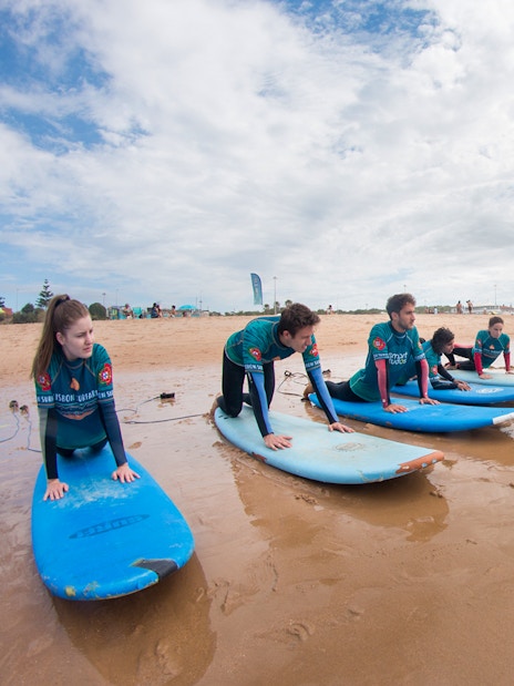 Group surf lesson on a sandy beach with instructor guiding participants on surfboards.