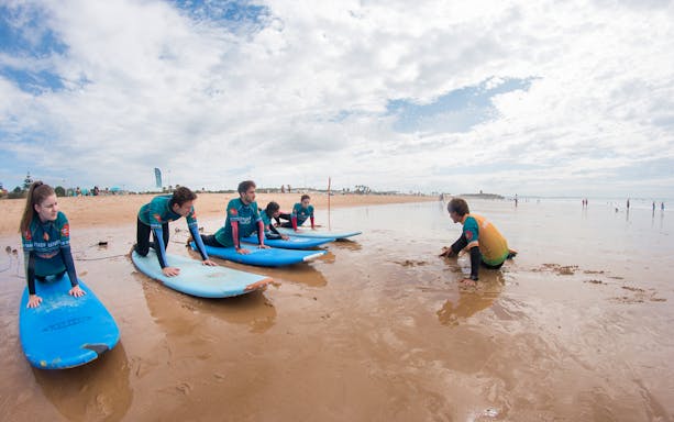 Group surf lesson on a sandy beach with instructor guiding participants on surfboards.
