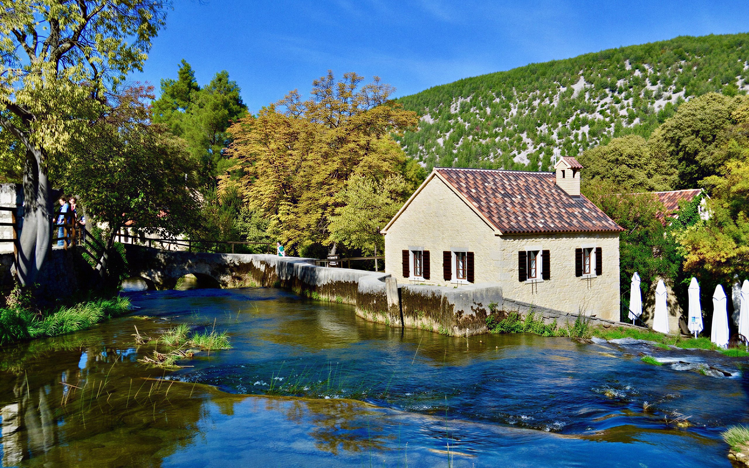 Old water mill by a stream in Krka National Park, surrounded by trees and hills.