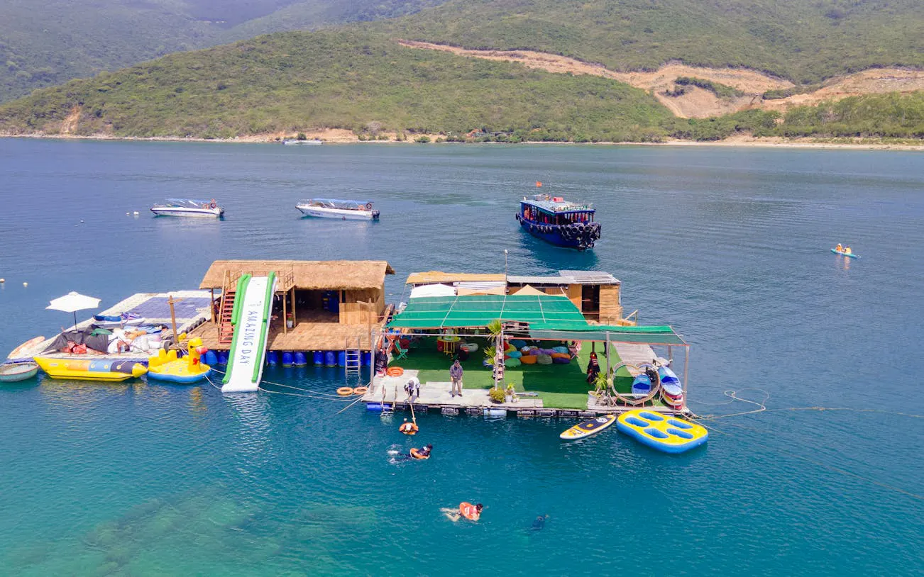 Floating platform with slide and boats at Green Oasis Beach, Nha Trang, with people snorkeling and paddleboarding.