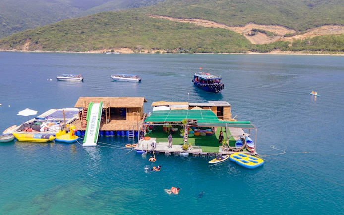 Floating platform with slide and boats at Green Oasis Beach, Nha Trang, with people snorkeling and paddleboarding.