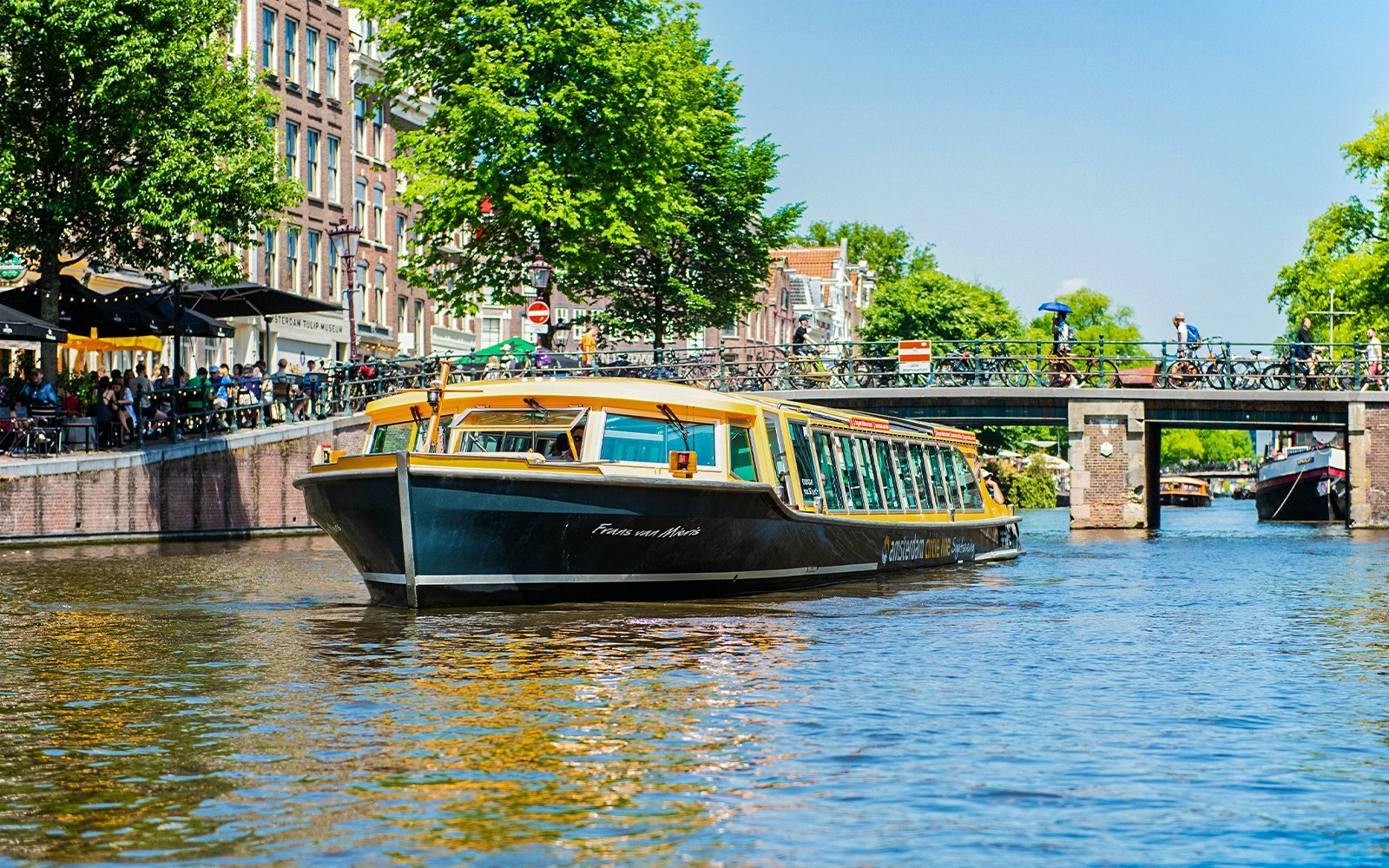 Boat cruising through Amsterdam canal under a bridge, daytime.