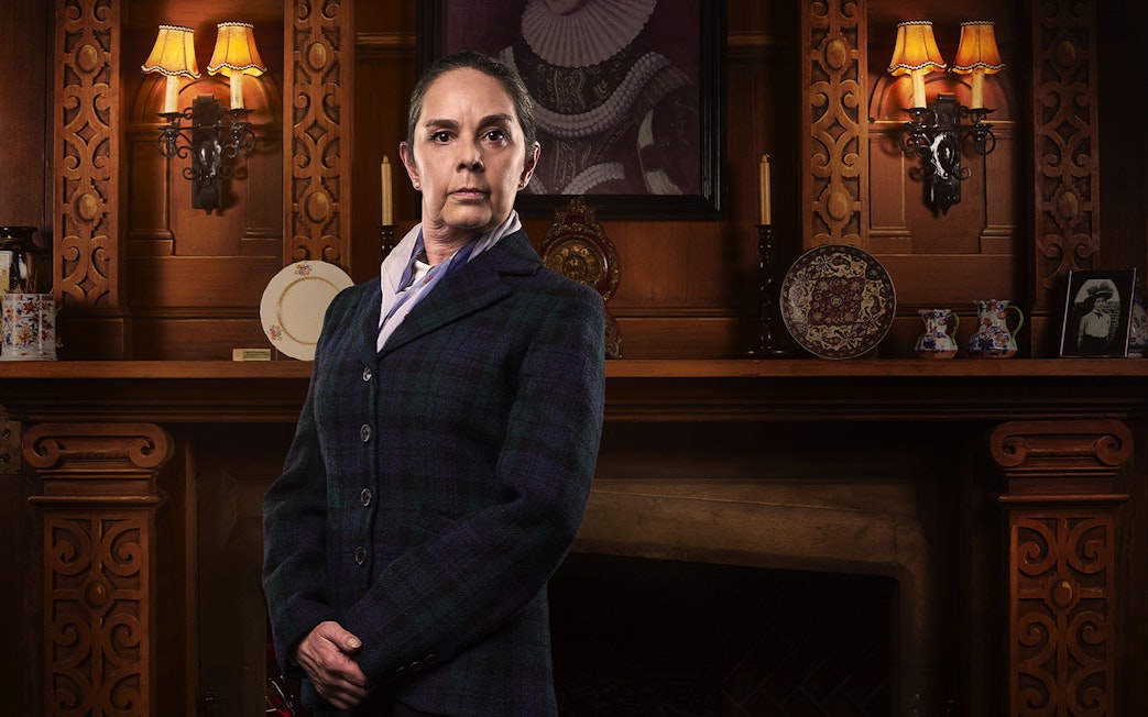 Person in period costume standing in front of ornate wooden fireplace, related to The Mousetrap.