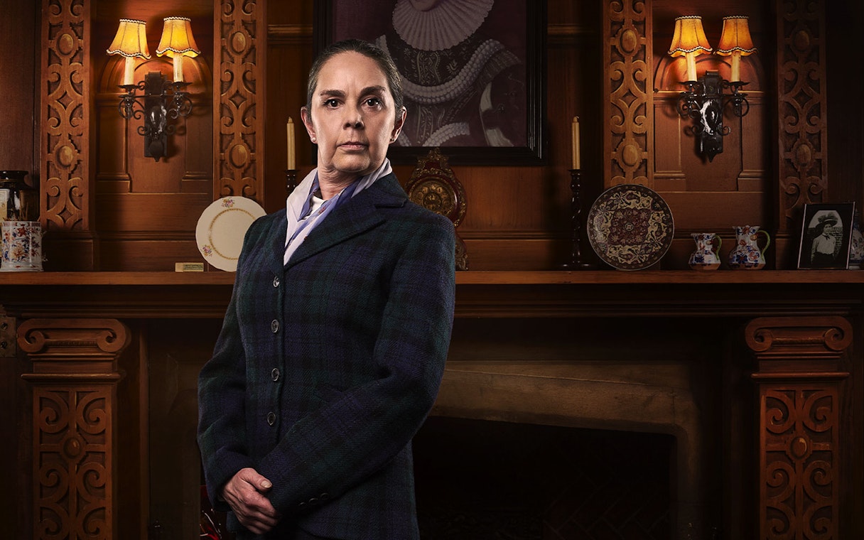 Person in period costume standing in front of ornate wooden fireplace, related to The Mousetrap.