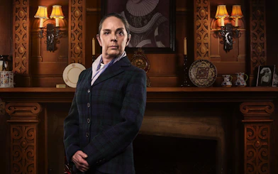 Person in period costume standing in front of ornate wooden fireplace, related to The Mousetrap.