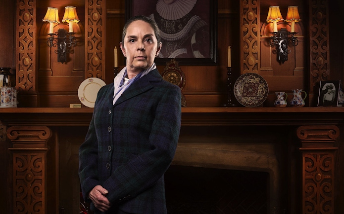 Person in period costume standing in front of ornate wooden fireplace, related to The Mousetrap.