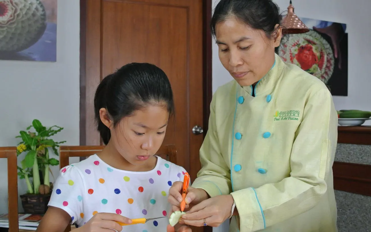Child learning fruit carving with a master carver in Thailand.