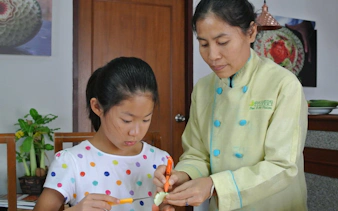Child learning fruit carving with a master carver in Thailand.