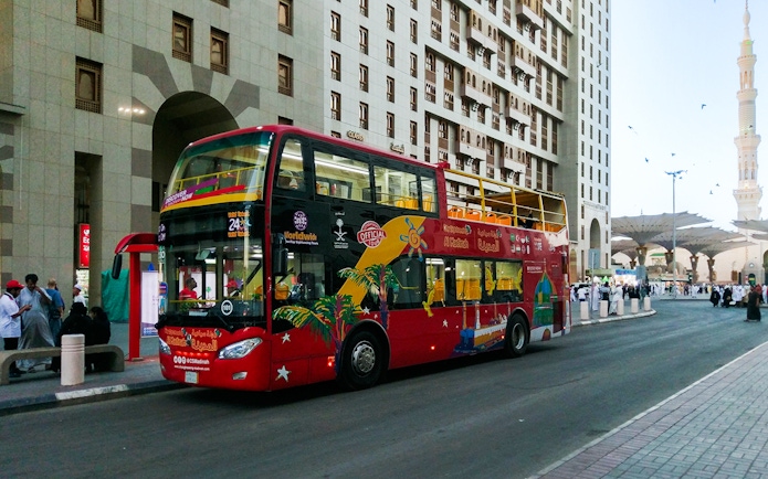 Red double-decker bus on Al Madinah Hop-On Hop-Off Tour near city buildings.