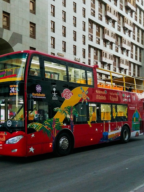 Red double-decker bus on Al Madinah Hop-On Hop-Off Tour near city buildings.