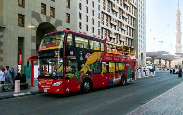 Red double-decker bus on Al Madinah Hop-On Hop-Off Tour near city buildings.