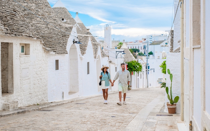 Tourist couple walking on cobblestone street in Alberobello, Puglia, with trulli houses.