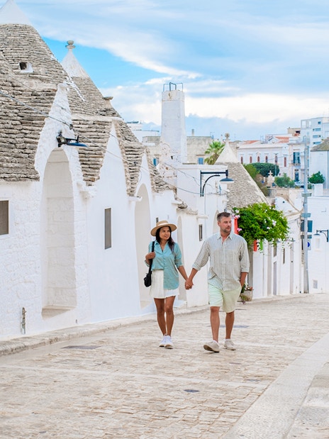 Tourist couple walking on cobblestone street in Alberobello, Puglia, with trulli houses.
