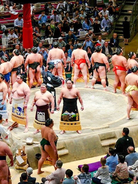 Sumo wrestlers in a Tokyo arena preparing for a match, surrounded by spectators.