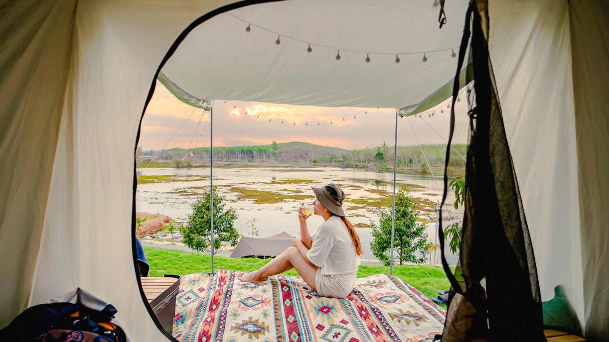 Solo female traveler relaxing in a tent overlooking a scenic lake at sunset.