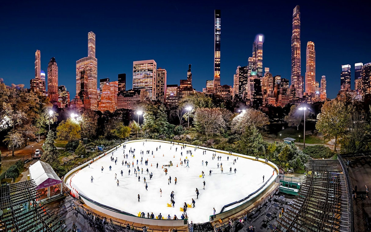 Wollman Rink in Central Park with skaters and New York City skyline at night.