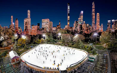Wollman Rink in Central Park with skaters and New York City skyline at night.