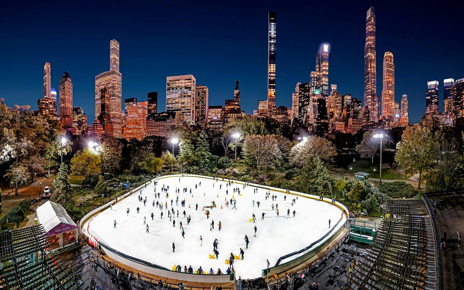 Wollman Rink in Central Park with skaters and New York City skyline at night.