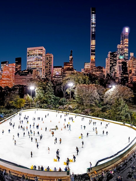 Wollman Rink in Central Park with skaters and New York City skyline at night.