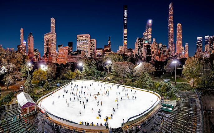 Wollman Rink in Central Park with skaters and New York City skyline at night.