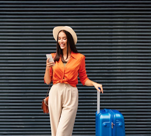 Female traveler with blue luggage checking phone in urban setting.