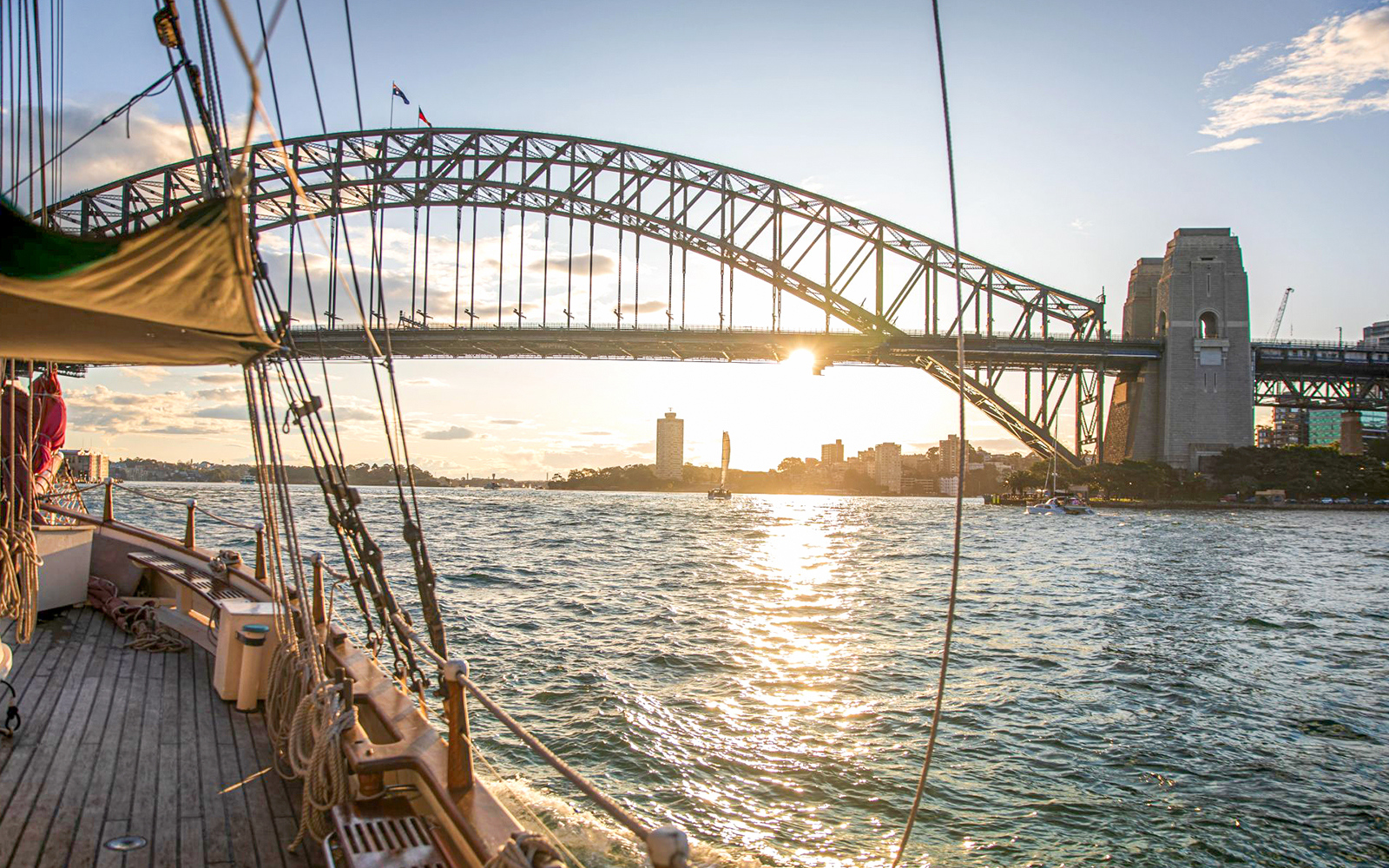 Tall ship sailing under Sydney Harbour Bridge at sunset.