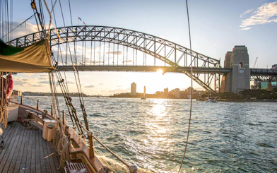 Tall ship sailing under Sydney Harbour Bridge at sunset.