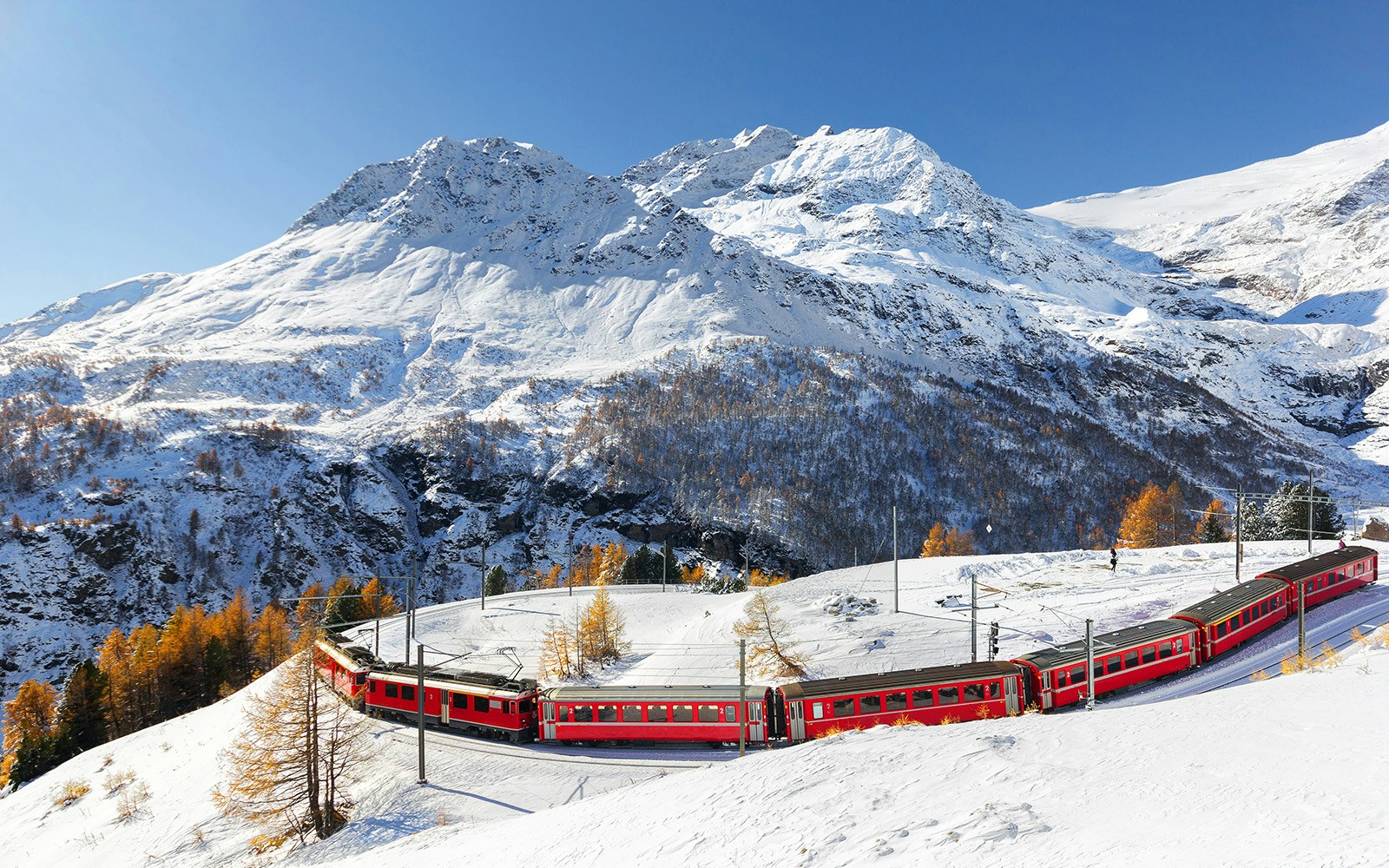 A red train is passing the train tracks with tight 180° curve at high Alp Ghe with the Piz Palu peak is at the background.