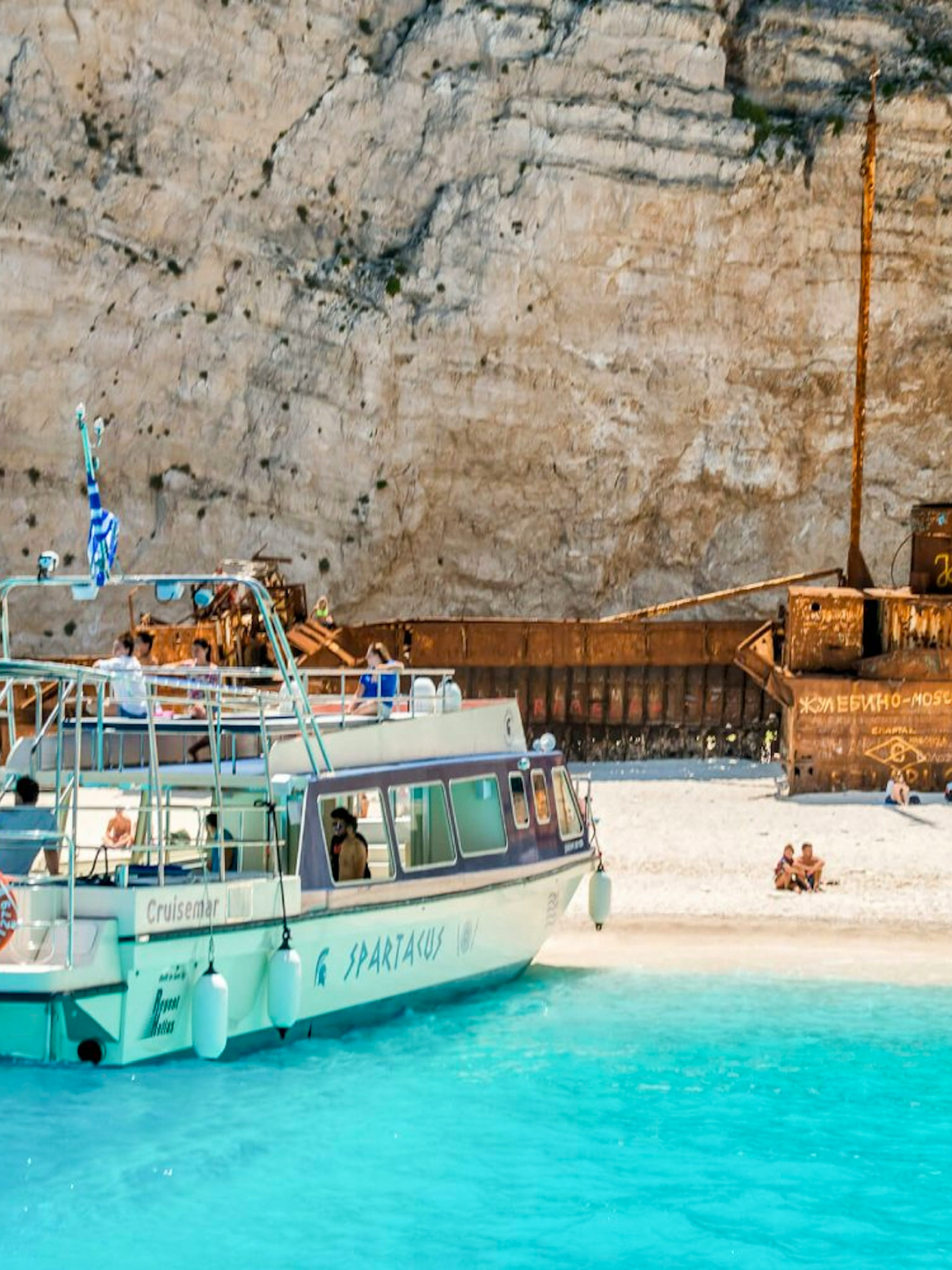 Glass bottom boat near shipwreck on Navagio Beach, Zakynthos, with tourists exploring the shore.