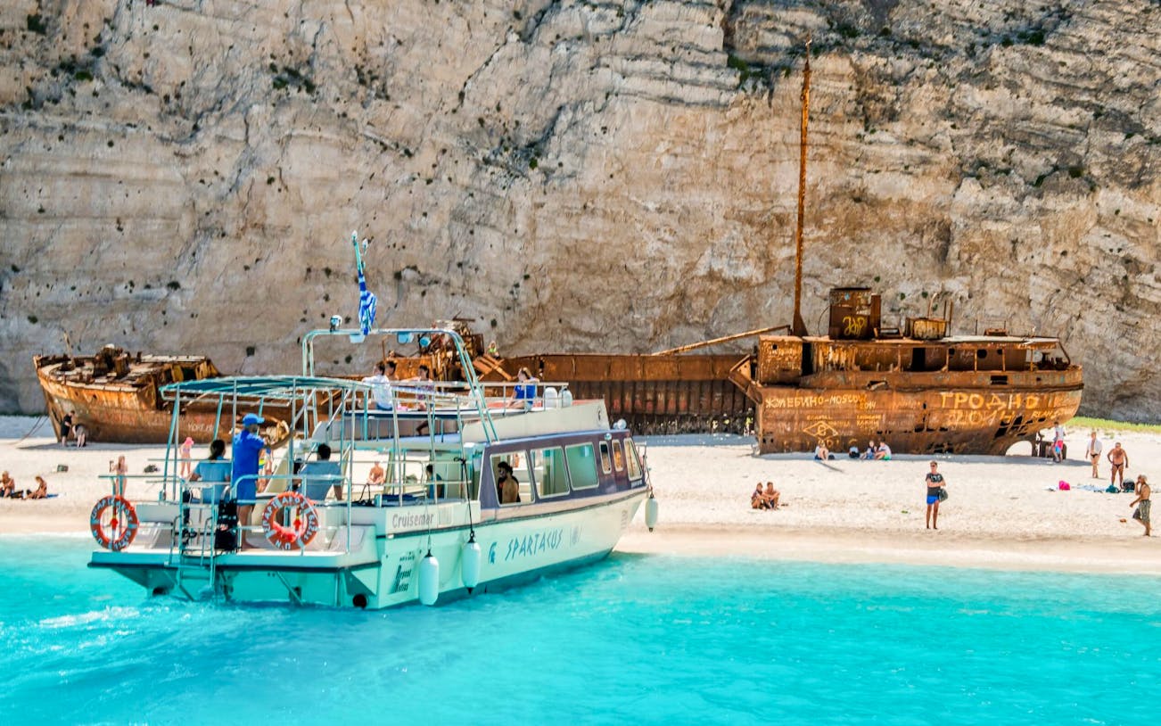 Glass bottom boat near shipwreck on Navagio Beach, Zakynthos, with tourists exploring the shore.
