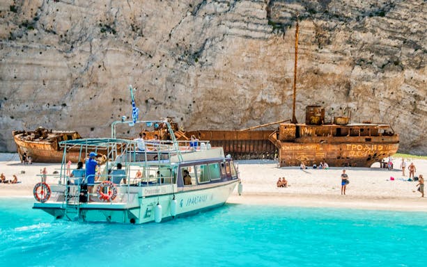 Glass bottom boat near shipwreck on Navagio Beach, Zakynthos, with tourists exploring the shore.