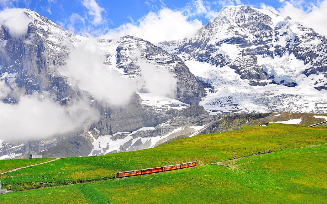 Train traveling through green fields with snow-capped mountains in Jungfraujoch, Switzerland.