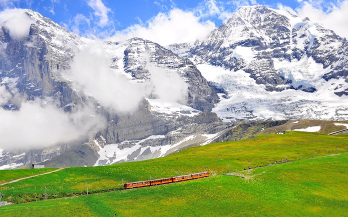 Train traveling through green fields with snow-capped mountains in Jungfraujoch, Switzerland.