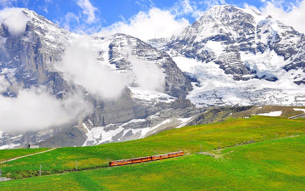 Train traveling through green fields with snow-capped mountains in Jungfraujoch, Switzerland.