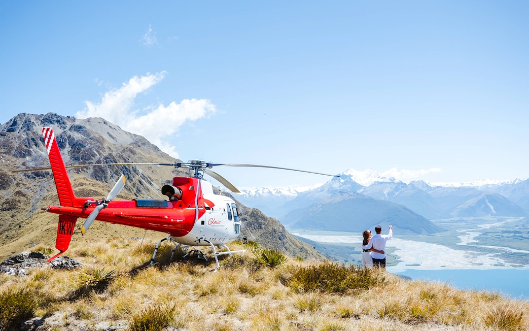 Couple by helicopter at Queenstown viewpoint overlooking mountains and river valley.