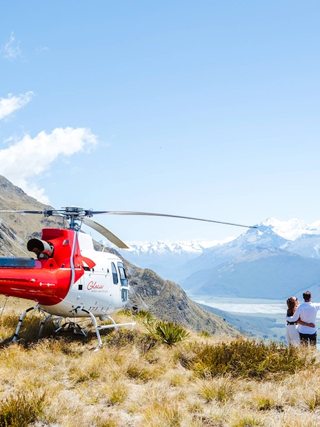 Couple by helicopter at Queenstown viewpoint overlooking mountains and river valley.