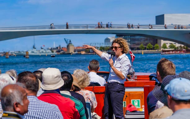 Tour guide speaking to passengers on a boat during a Copenhagen canal tour with a bridge in the background.