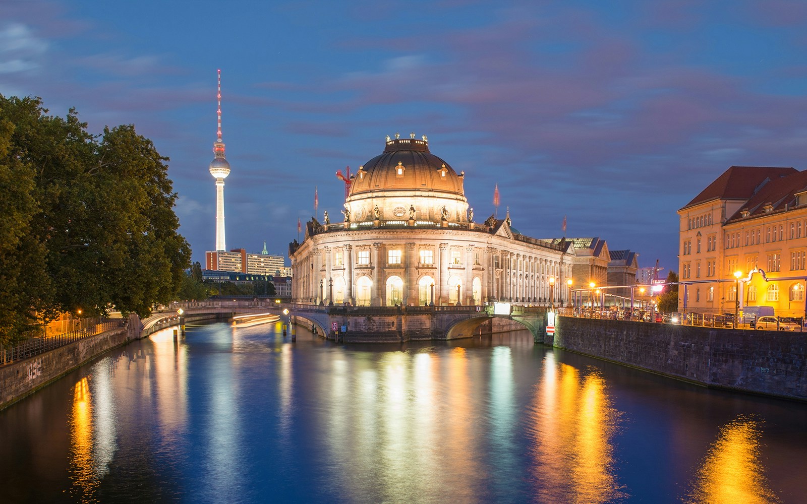 Das Bode-Museum in Berlin bei Nacht, beleuchtet und reflektiert im Wasser der Spree, mit dem Fernsehturm im Hintergrund.