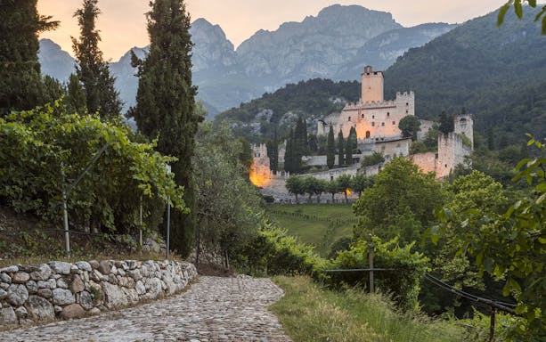 Avio Castle in Italy surrounded by lush greenery and mountains at sunset.