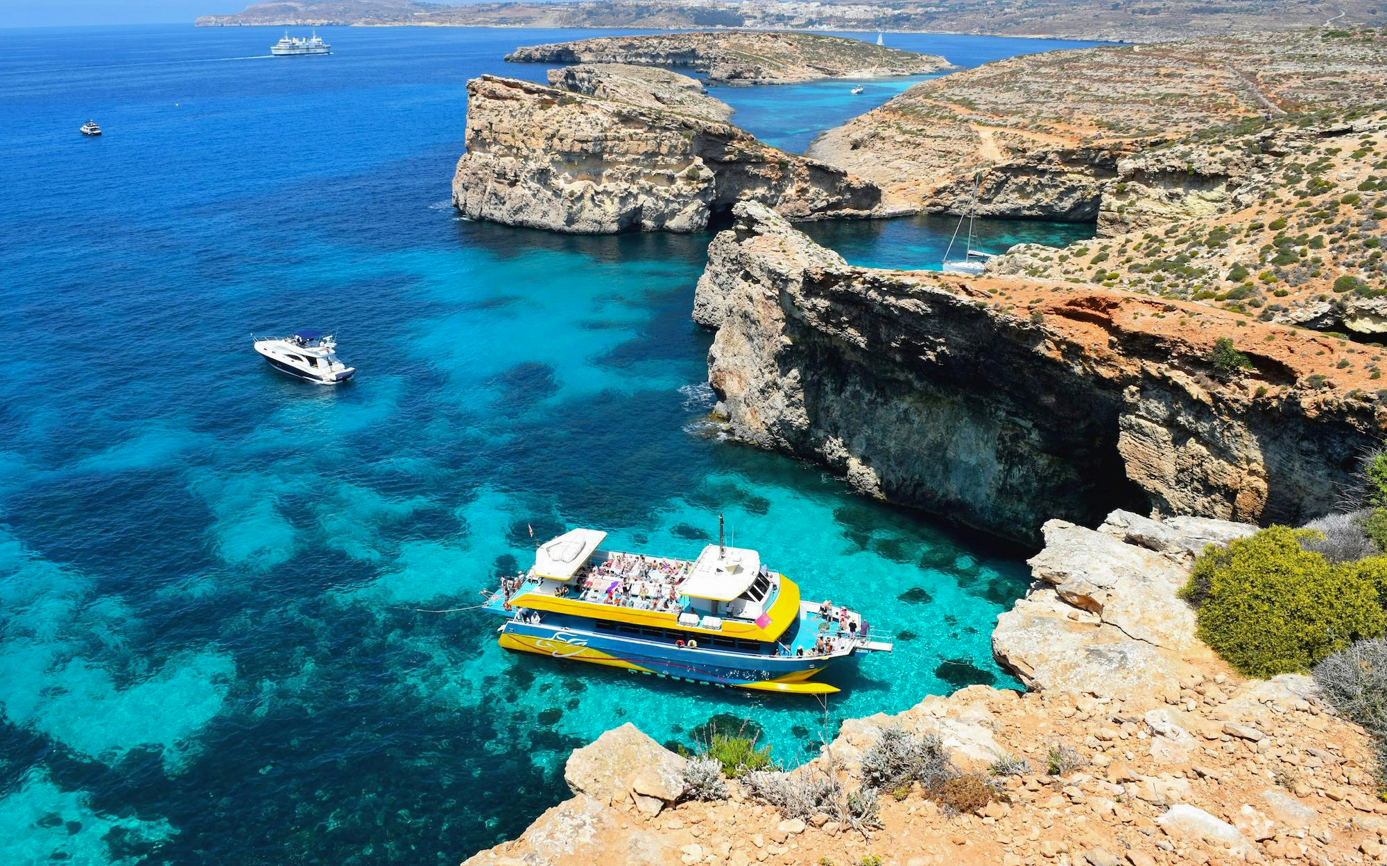 Aerial view of a boat in the Blue Lagoon, Malta, surrounded by clear turquoise waters and rocky cliffs.