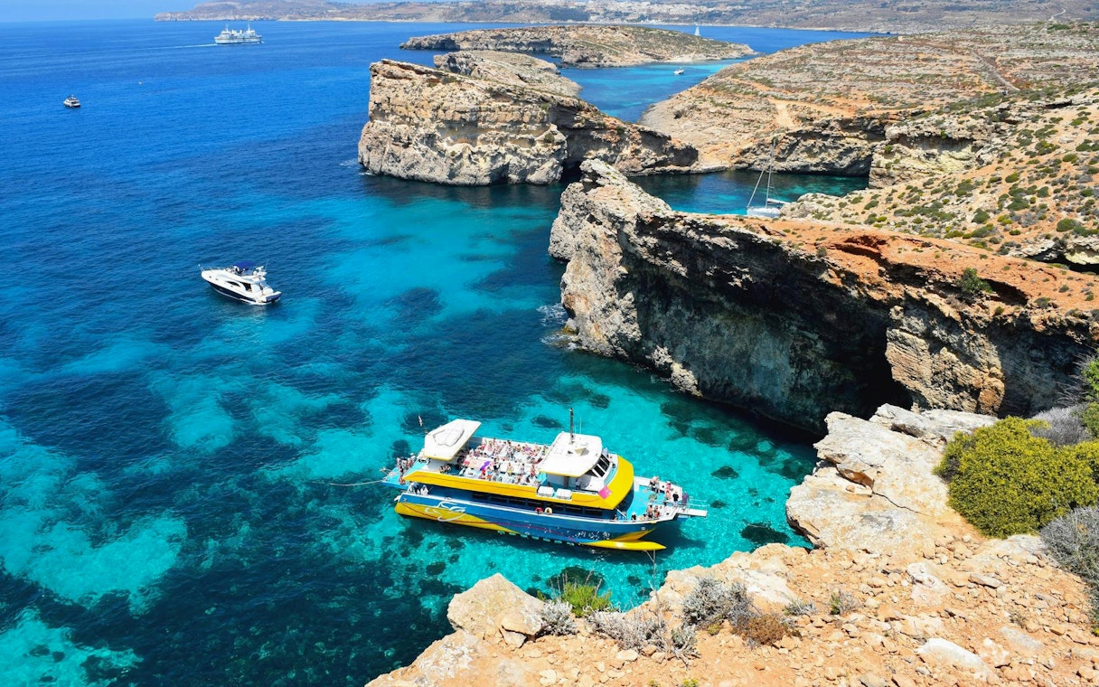 Aerial view of a boat in the Blue Lagoon, Malta, surrounded by clear turquoise waters and rocky cliffs.