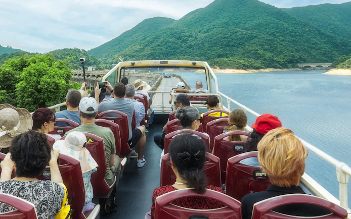 Passengers on an open-top bus tour in Hong Kong with views of lush hills and a waterway.