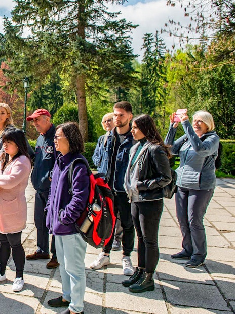 Group of tourists listening to a guide at Bran Castle, surrounded by lush greenery.