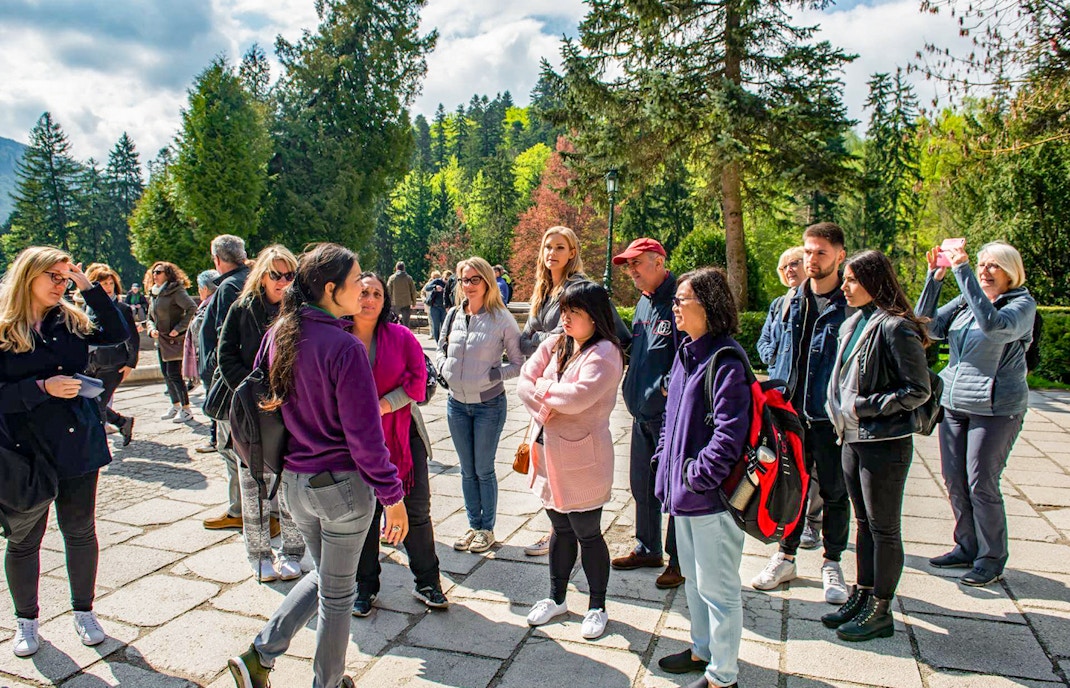 Group of tourists listening to a guide at Bran Castle, surrounded by lush greenery.