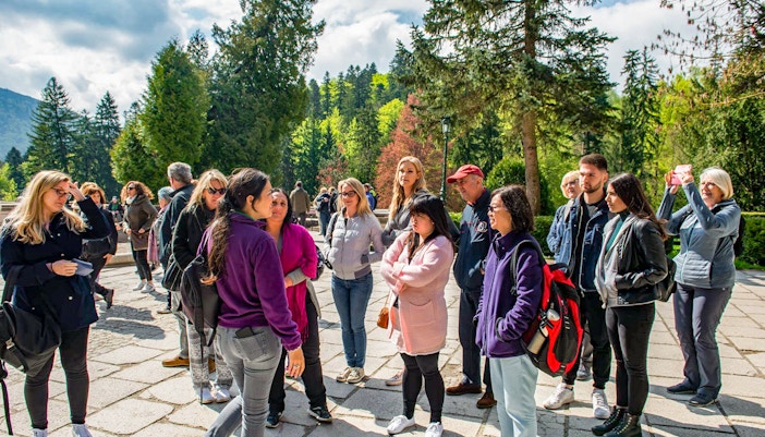 Group of tourists listening to a guide at Bran Castle, surrounded by lush greenery.