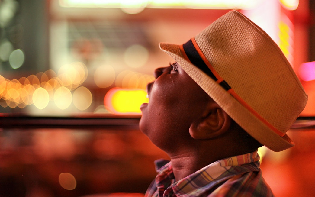 Child wearing a hat looking out of a bus window at night city lights.