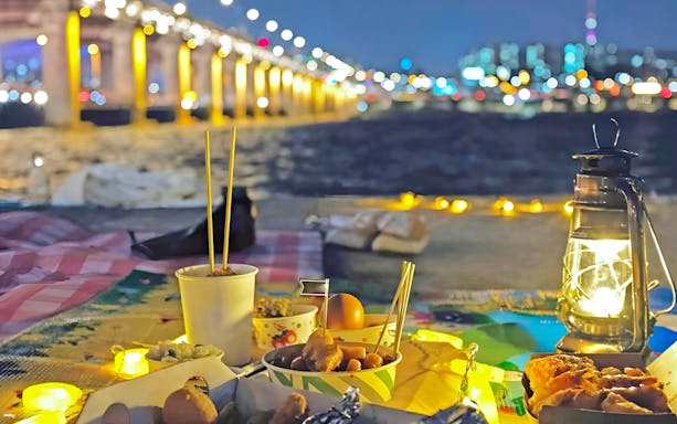 Night picnic setup by Han River with food, lantern, and bridge lights in Seoul.