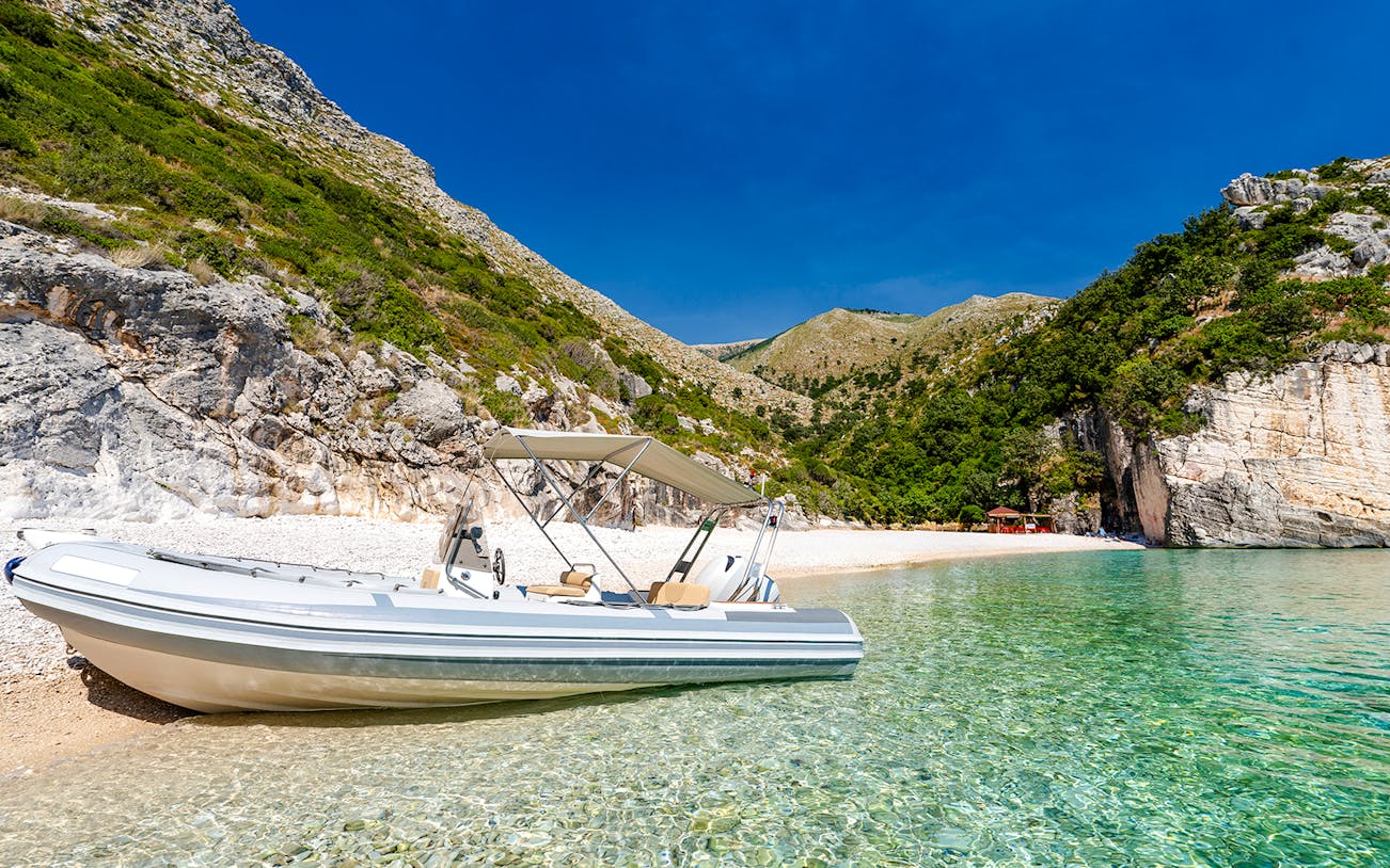 Boat on clear waters at Grama Bay, Albania, with rocky cliffs and lush greenery in the background.